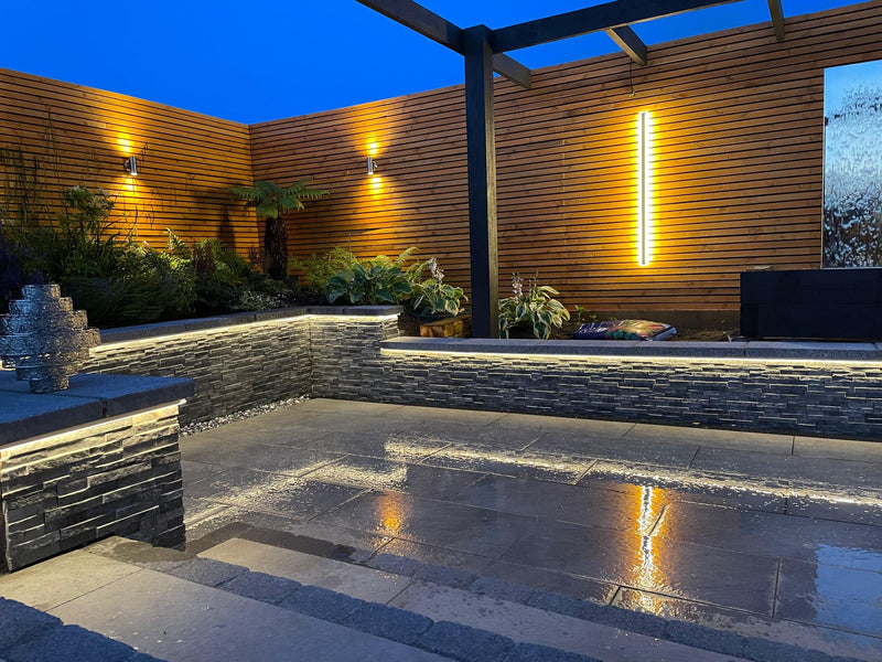 Outdoor patio area with stone seating, plants, and wall lights at night.